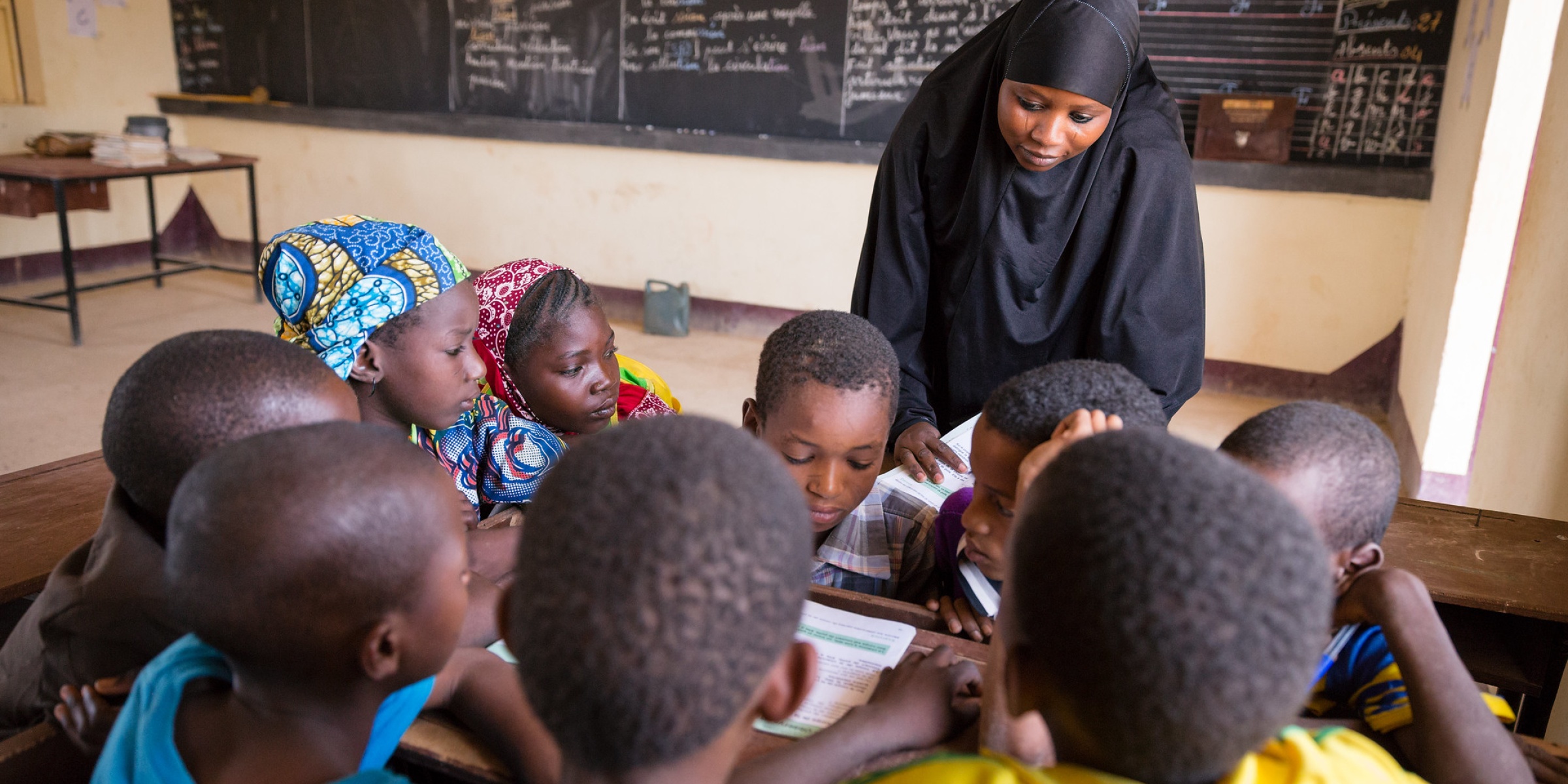 Grade three students share a textbook with the teacher looking on.